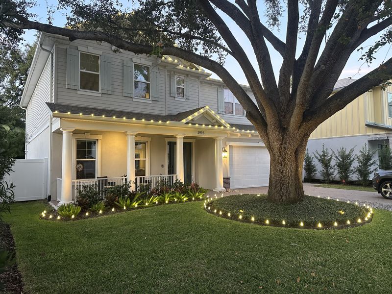 Two-story home with warm white roofline lights and a lit ring around a large oak tree, installed by Exterior Holiday Lights